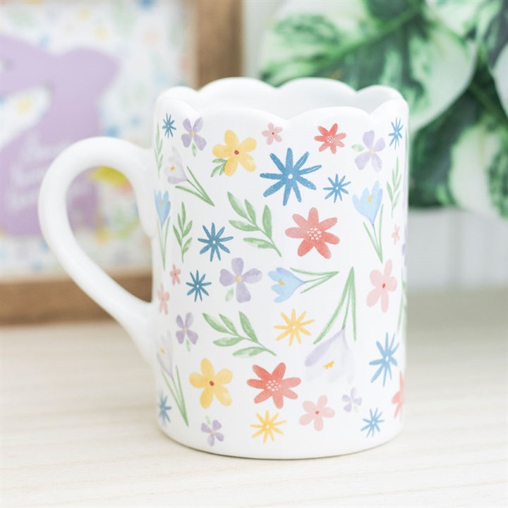 A white ceramic mug decorated with a colorful array of pastel flowers and green leaves. The mug features a unique scalloped rim and curved handle, creating a soft and cheerful springtime aesthetic.
