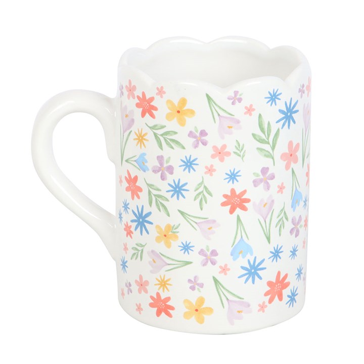 A white ceramic mug decorated with a colorful array of pastel flowers and green leaves. The mug features a unique scalloped rim and curved handle, creating a soft and cheerful springtime aesthetic.