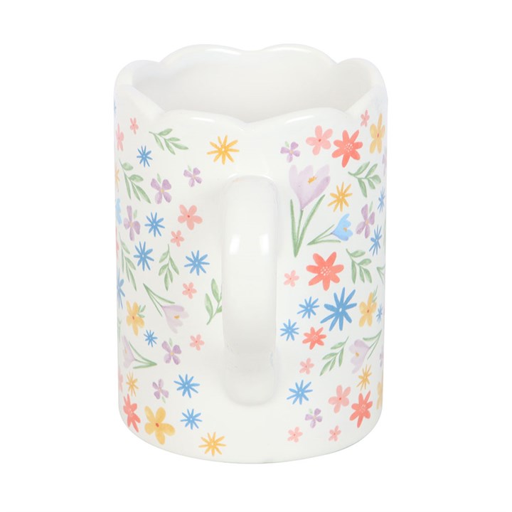 A white ceramic mug decorated with a colorful array of pastel flowers and green leaves. The mug features a unique scalloped rim and curved handle, creating a soft and cheerful springtime aesthetic.