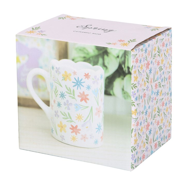 A white ceramic mug decorated with a colorful array of pastel flowers and green leaves. The mug features a unique scalloped rim and curved handle, creating a soft and cheerful springtime aesthetic.