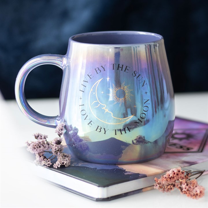 A glossy, iridescent mug in purple and blue tones featuring a celestial design with the phrase “Live by the Sun, Love by the Moon” and artwork of the sun, moon, and stars. The mug sits on top of a closed book with dried flowers, creating a dreamy, mystical atmosphere.