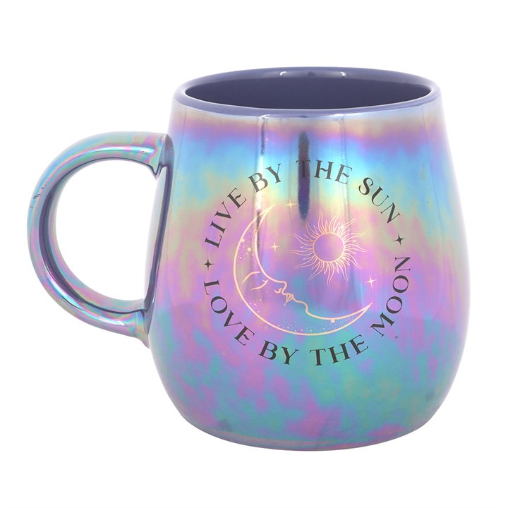A glossy, iridescent mug in purple and blue tones featuring a celestial design with the phrase “Live by the Sun, Love by the Moon” and artwork of the sun, moon, and stars. The mug sits on top of a closed book with dried flowers, creating a dreamy, mystical atmosphere.