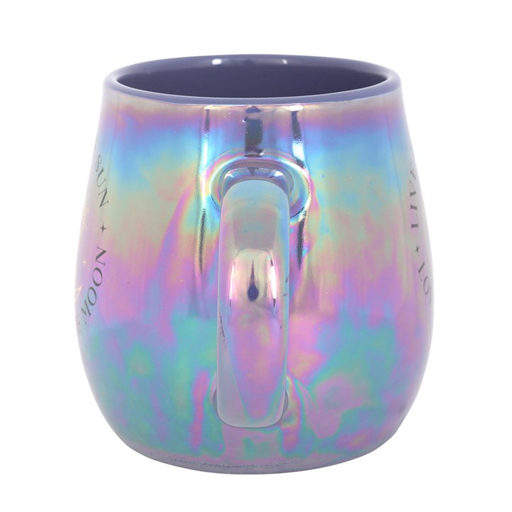 A glossy, iridescent mug in purple and blue tones featuring a celestial design with the phrase “Live by the Sun, Love by the Moon” and artwork of the sun, moon, and stars. The mug sits on top of a closed book with dried flowers, creating a dreamy, mystical atmosphere.