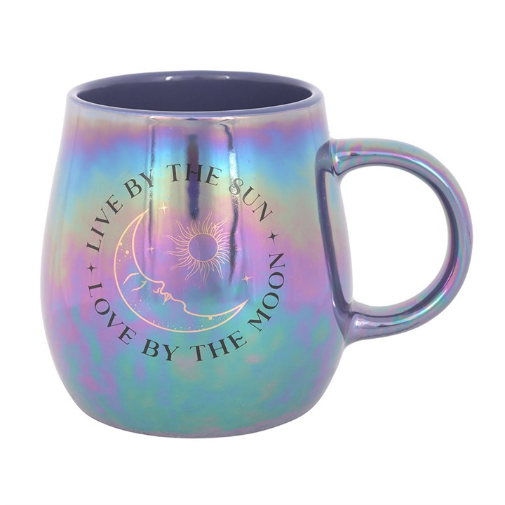 A glossy, iridescent mug in purple and blue tones featuring a celestial design with the phrase “Live by the Sun, Love by the Moon” and artwork of the sun, moon, and stars. The mug sits on top of a closed book with dried flowers, creating a dreamy, mystical atmosphere.
