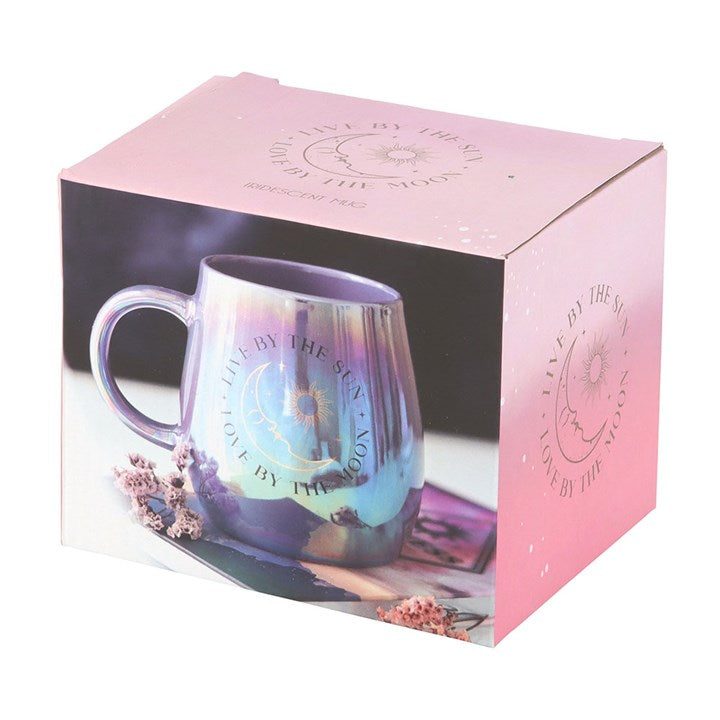 A glossy, iridescent mug in purple and blue tones featuring a celestial design with the phrase “Live by the Sun, Love by the Moon” and artwork of the sun, moon, and stars. The mug sits on top of a closed book with dried flowers, creating a dreamy, mystical atmosphere.