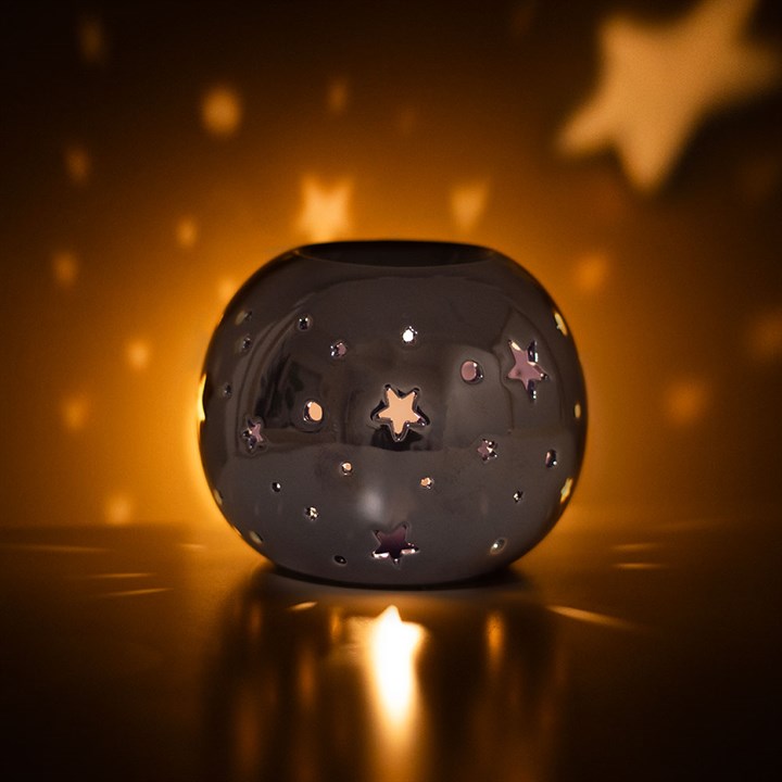 A round, purple iridescent oil burner with star and circle cut-outs that allow light to shine through. It sits beside a small essential oil bottle on a smooth surface, creating a dreamy and calming atmosphere ideal for relaxation.