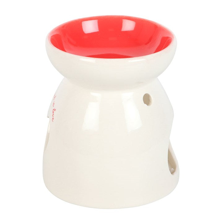Ceramic wax melt burner with 'All You Need Is Love' text, featuring a heart-shaped cutout glowing with a tealight candle and a red interior bowl. Perfect for home fragrance lovers to create a cozy and romantic ambiance