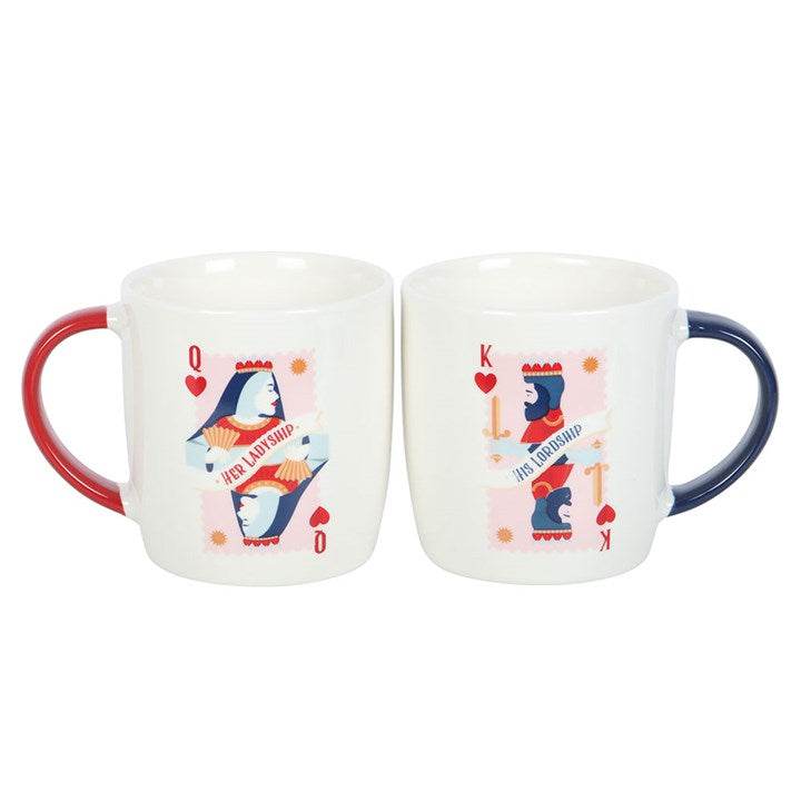 A pair of white ceramic mugs featuring playing card-inspired designs labeled “His Lordship” and “Her Ladyship.” One has a navy handle with a king of hearts motif, while the other has a red handle with a queen of hearts design. The mugs are displayed on a round white tray against a cozy navy background.