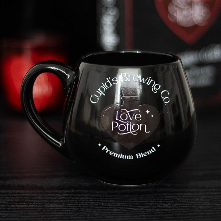Black ceramic Love Potion heat change mug featuring 'Cupid's Brewing Co.' design with a heart-shaped potion bottle graphic that changes color when filled with a hot beverage.