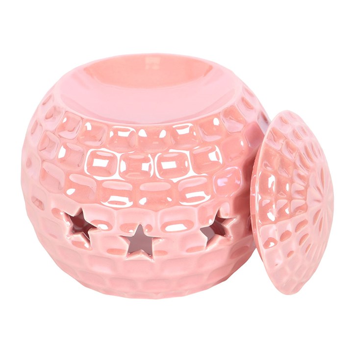 A glossy pink ceramic oil burner shaped like a disco ball, featuring star-shaped cut-outs. The burner sits atop a closed book, surrounded by soft, decorative elements, adding a fun and nostalgic touch to the space.