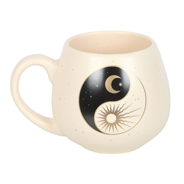 A cream-colored ceramic mug with a rounded shape, featuring a yin and yang design that includes a crescent moon with stars on one side and a radiant sun on the other. The mug rests on a book, creating a calm and serene atmosphere.