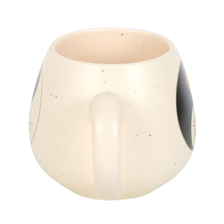 A cream-colored ceramic mug with a rounded shape, featuring a yin and yang design that includes a crescent moon with stars on one side and a radiant sun on the other. The mug rests on a book, creating a calm and serene atmosphere.