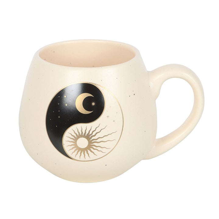 A cream-colored ceramic mug with a rounded shape, featuring a yin and yang design that includes a crescent moon with stars on one side and a radiant sun on the other. The mug rests on a book, creating a calm and serene atmosphere.