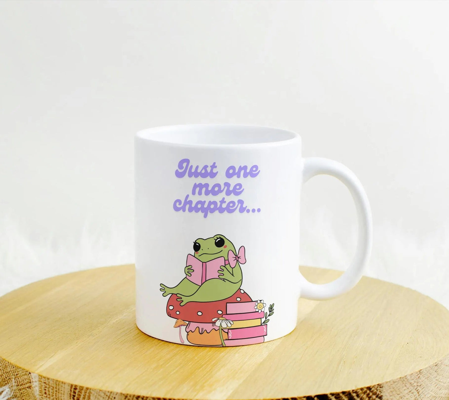 A white ceramic mug featuring a cute illustration of a green frog with a pink bow, sitting on a red mushroom and holding a book. Below the frog are stacked pink books with flowers, and above it, the text reads "Just one more chapter..." in purple lettering.