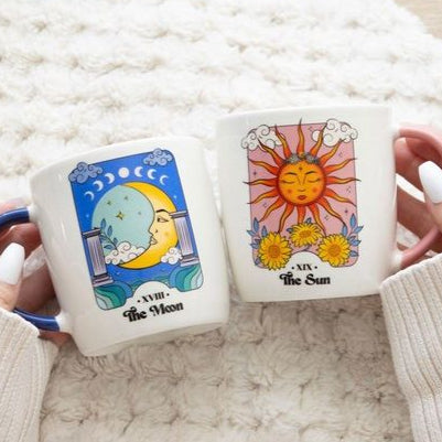 A set of two ceramic mugs inspired by tarot cards. One mug features the Sun card with a bright sun face, pink handle, and floral accents. The other displays the Moon card with a crescent moon and serene night sky, paired with a blue handle. Both have a white base and colourful celestial artwork.