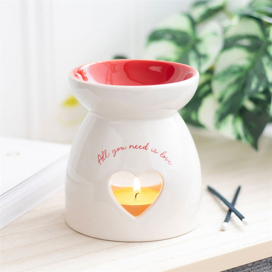 Ceramic wax melt burner with 'All You Need Is Love' text, featuring a heart-shaped cutout glowing with a tealight candle and a red interior bowl. Perfect for home fragrance lovers to create a cozy and romantic ambiance