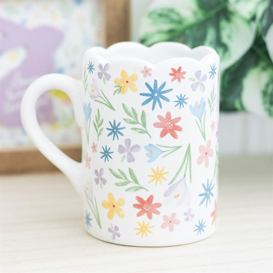 A white ceramic mug decorated with a colorful array of pastel flowers and green leaves. The mug features a unique scalloped rim and curved handle, creating a soft and cheerful springtime aesthetic.