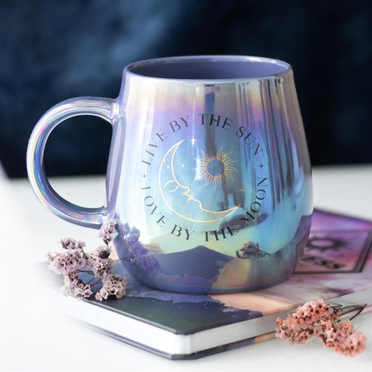 A glossy, iridescent mug in purple and blue tones featuring a celestial design with the phrase “Live by the Sun, Love by the Moon” and artwork of the sun, moon, and stars. The mug sits on top of a closed book with dried flowers, creating a dreamy, mystical atmosphere.