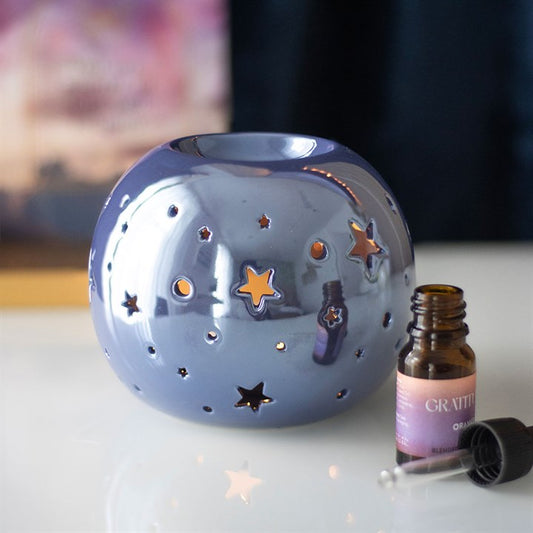 A round, purple iridescent oil burner with star and circle cut-outs that allow light to shine through. It sits beside a small essential oil bottle on a smooth surface, creating a dreamy and calming atmosphere ideal for relaxation.