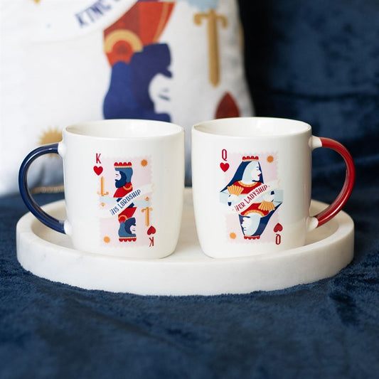 A pair of white ceramic mugs featuring playing card-inspired designs labeled “His Lordship” and “Her Ladyship.” One has a navy handle with a king of hearts motif, while the other has a red handle with a queen of hearts design. The mugs are displayed on a round white tray against a cozy navy background.