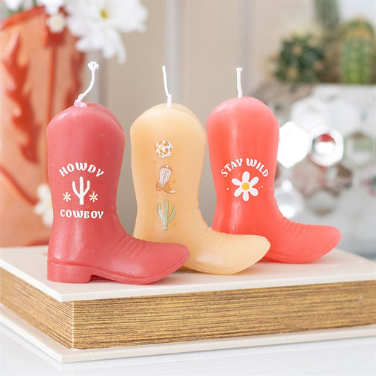 A set of three unscented decorative candles shaped like cowboy boots in shades of pink and peach. Each boot features Western-themed designs and phrases like "Howdy Cowboy" and "Stay Wild." Displayed on a wooden surface with soft background decor, adding a playful country charm.