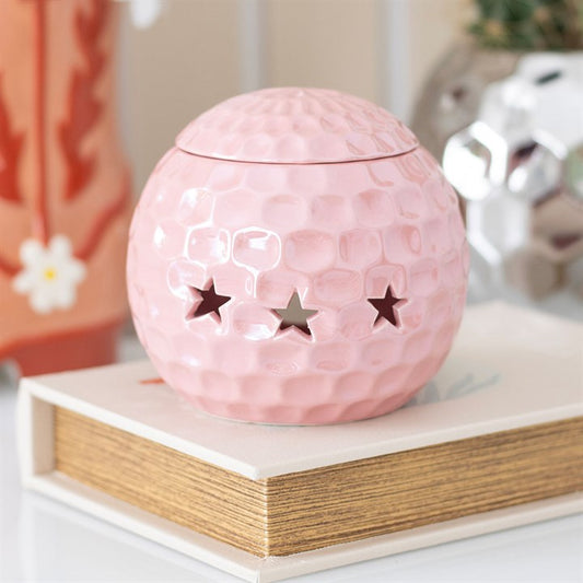 A glossy pink ceramic oil burner shaped like a disco ball, featuring star-shaped cut-outs. The burner sits atop a closed book, surrounded by soft, decorative elements, adding a fun and nostalgic touch to the space.