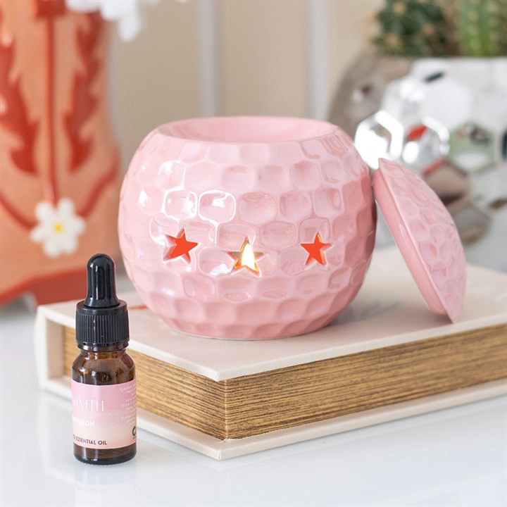 A glossy pink ceramic oil burner shaped like a disco ball, featuring star-shaped cut-outs. The burner sits atop a closed book, surrounded by soft, decorative elements, adding a fun and nostalgic touch to the space.