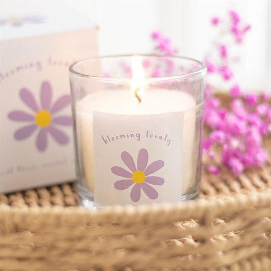A clear glass candle with a lit flame, featuring a purple and yellow daisy illustration and the phrase "blooming lovely" on the label. The candle is placed on a woven surface with a matching box and soft pink flowers in the background, creating a warm and cheerful vibe.