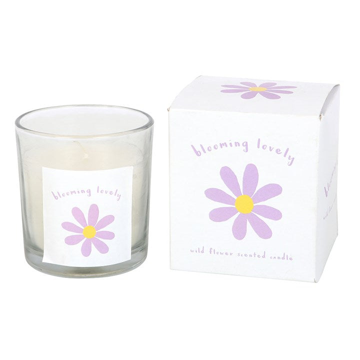 A clear glass candle with a lit flame, featuring a purple and yellow daisy illustration and the phrase "blooming lovely" on the label. The candle is placed on a woven surface with a matching box and soft pink flowers in the background, creating a warm and cheerful vibe.