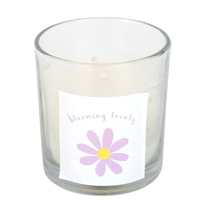 A clear glass candle with a lit flame, featuring a purple and yellow daisy illustration and the phrase "blooming lovely" on the label. The candle is placed on a woven surface with a matching box and soft pink flowers in the background, creating a warm and cheerful vibe.