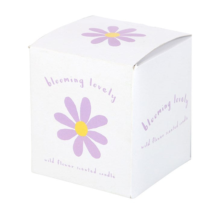 A clear glass candle with a lit flame, featuring a purple and yellow daisy illustration and the phrase "blooming lovely" on the label. The candle is placed on a woven surface with a matching box and soft pink flowers in the background, creating a warm and cheerful vibe.