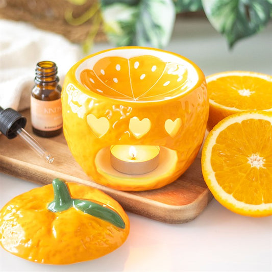 A ceramic orange-shaped oil burner with a glossy finish, featuring cut-out heart shapes that allow the light from a tealight candle to shine through.