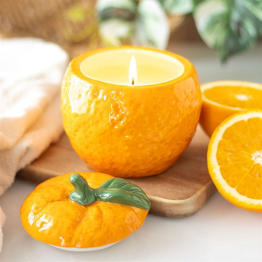 A beautifully designed orange-shaped scented candle housed in a glossy ceramic jar with a removable lid, featuring a green leaf accent. 