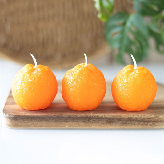 A set of three unscented, orange-shaped candles with a textured surface, placed on a wooden board. The candles have a bright orange hue and white wicks, resembling real oranges. 