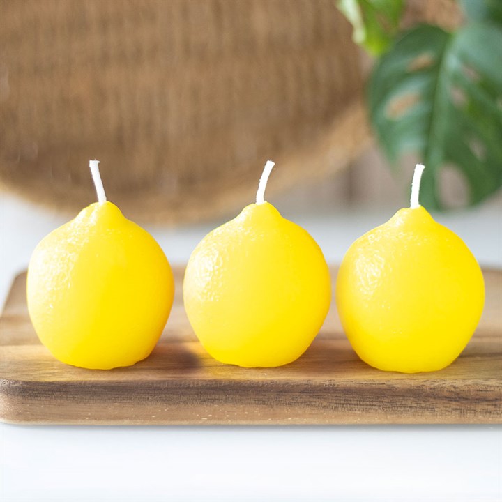 A set of three unscented, lemon-shaped candles with a smooth, glossy texture, placed on a wooden board. The candles have a bright yellow hue and white wicks, resembling real lemons