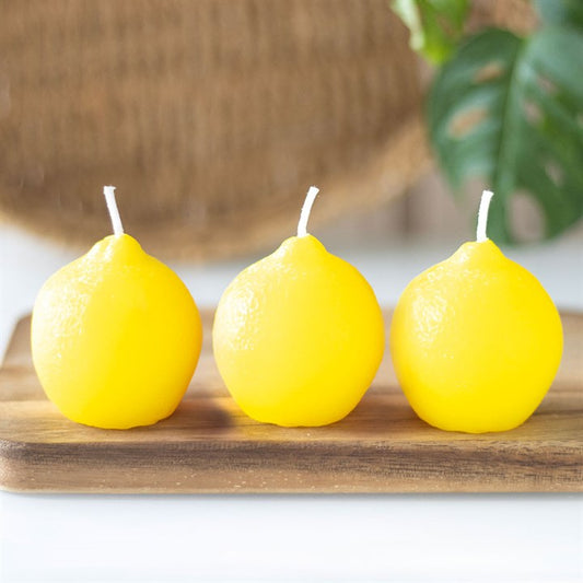 A set of three unscented, lemon-shaped candles with a smooth, glossy texture, placed on a wooden board. The candles have a bright yellow hue and white wicks, resembling real lemons
