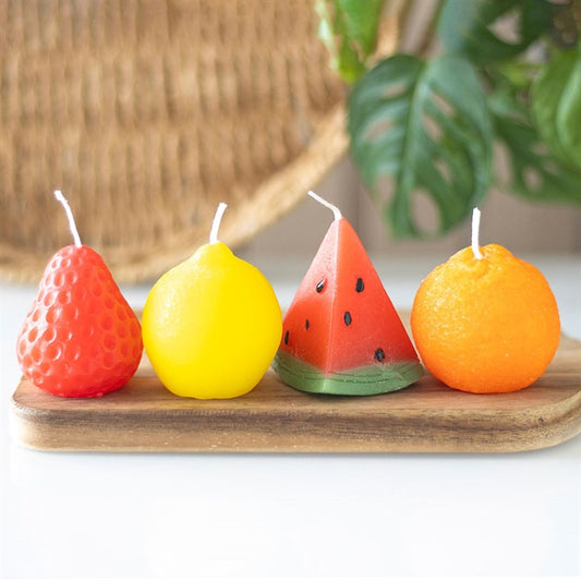 A set of four unscented, fruit-shaped candles—strawberry, lemon, watermelon, and orange—arranged on a wooden board. Each candle has a realistic texture and vibrant color, creating a fun and decorative display. 