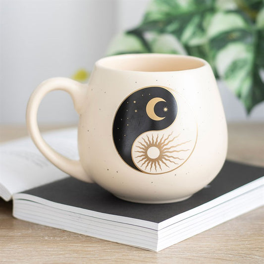 A cream-colored ceramic mug with a rounded shape, featuring a yin and yang design that includes a crescent moon with stars on one side and a radiant sun on the other. The mug rests on a book, creating a calm and serene atmosphere.