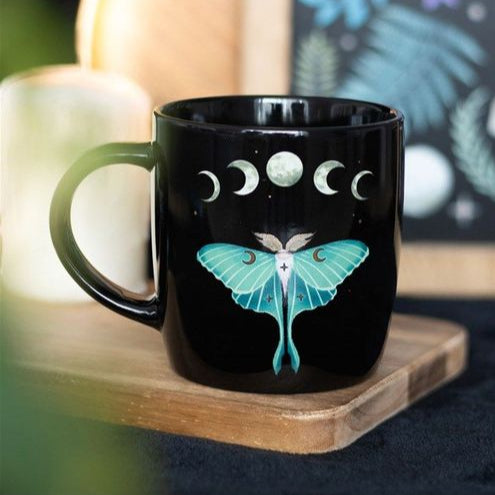 A glossy black ceramic mug featuring a detailed illustration of a teal luna moth, surrounded by moon phases, stars, and floral accents. The design evokes a mystical, nature-inspired aesthetic.